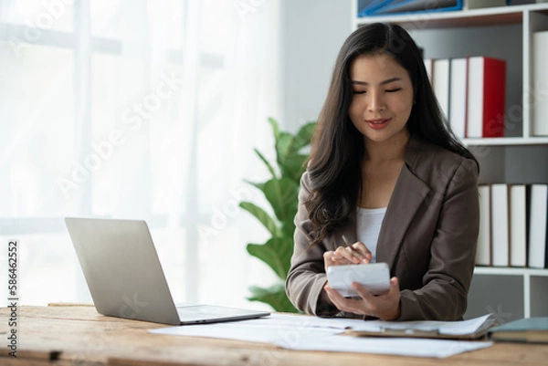 Fototapeta Asian businesswoman recording data using desktop computer keyboard and calculator to analyze marketing strategy and real estate data reduce corporate taxes for clients. Accounting and Tax Concepts