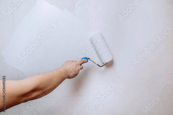 Fototapeta A man's hand holding a paint roller and priming the wall in white close-up. The concept of renovation in the apartment.