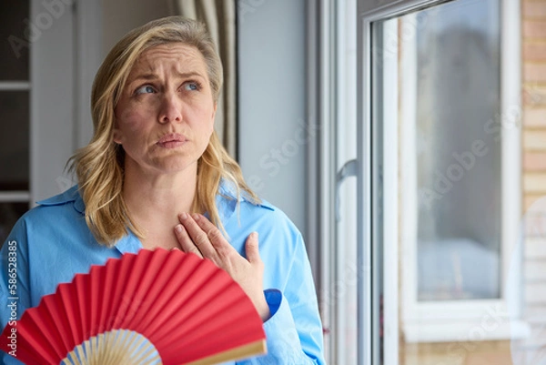 Obraz Menopausal Mature Woman Having Hot Flush At Home Cooling Herself With Handheld Paper Fan