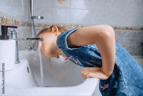 Fototapeta A little girl in a denim dress with a print leaned on a white ceramic sink to drink water from the tap in the bathroom.