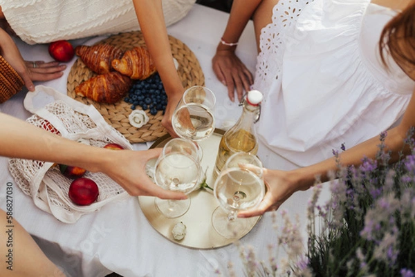 Fototapeta Friendship. Leisure at nature. Group of happy girlfriends clinking glasses with wine at picnic on lavender field. Delicious picnic with croissants, berries and wine.