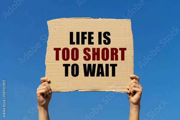 Fototapeta Life is too short to wait text on box paper held by 2 hands with isolated blue sky background. This message board can be used as business concept about starting doing something you would like to do no