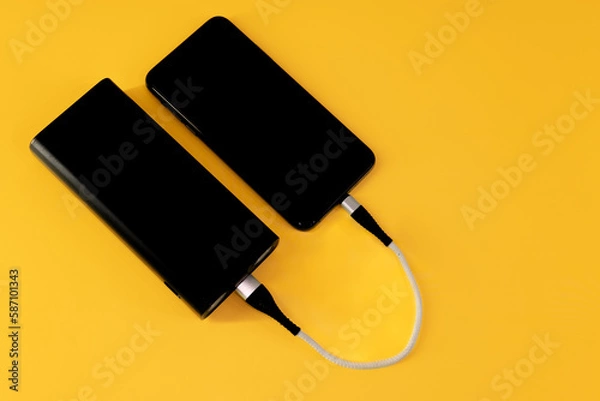 Obraz Mobile phone charges from black portable power bank with white cable on yellow background