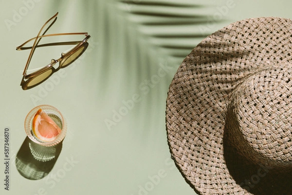 Fototapeta Summer flat lay with straw hat, sunglasses and glass of water with grapefruit slices on green background with palm leaf shadow, sun and sunlight. Vacation, holiday, summer creative minimal concept.