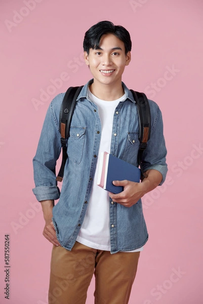 Fototapeta Smart guy student with backpack and bunch of books smiling at camera, copy space for advertisement over pink background