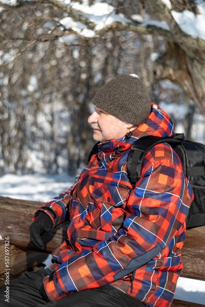 Fototapeta A man in a red plaid jacket and a gray hat with a backpack over his shoulders sits on a bench in the park