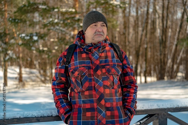 Fototapeta A man in a red plaid jacket and a gray hat is standing on the veranda against the backdrop of pine trees.