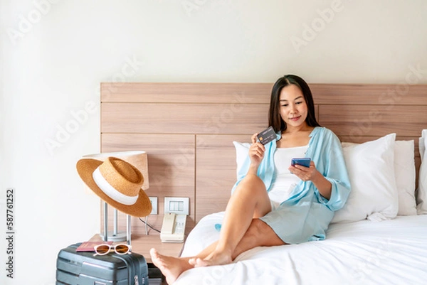 Fototapeta Happy traveler Asian woman holding credit card while using smartphone with travel accessories on bed. Online shopping and travel, cashless society, technology and lifestyle concept. Copy space