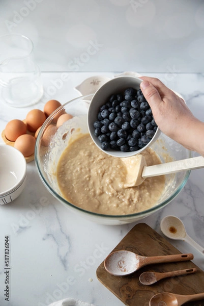 Fototapeta Glass mixing bowl with various baking ingredients around it. Ingredient is poured into bowl and mixed by hand.