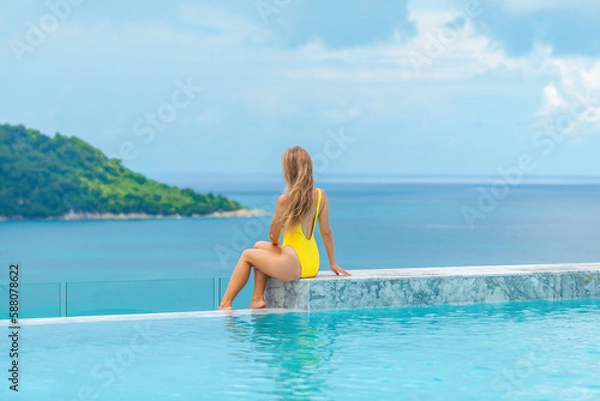 Fototapeta Back view of young woman in yellow swimwear relaxes in infinity pool at a luxury spa resort, enjoying the stunning sea view. A perfect summer vacation getaway.