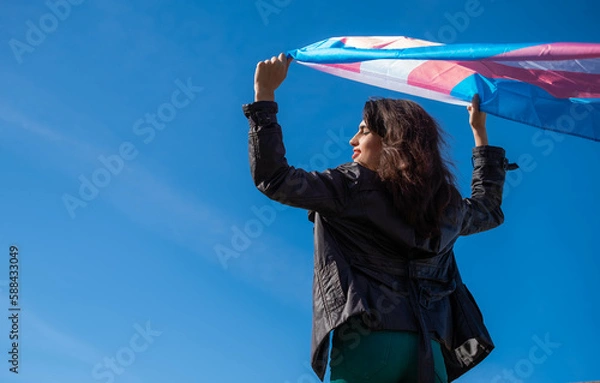 Fototapeta Transgender woman raising the trans flag in flight with a clear sky in the background. LGTBIQ+ activism