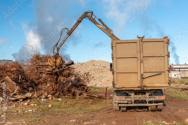 Obraz A grapple truck loads scrap industrial metal for recycling. Manipulator with a hydraulic magnetic crab against the sky. Crane garbage truck loading of industrial waste.