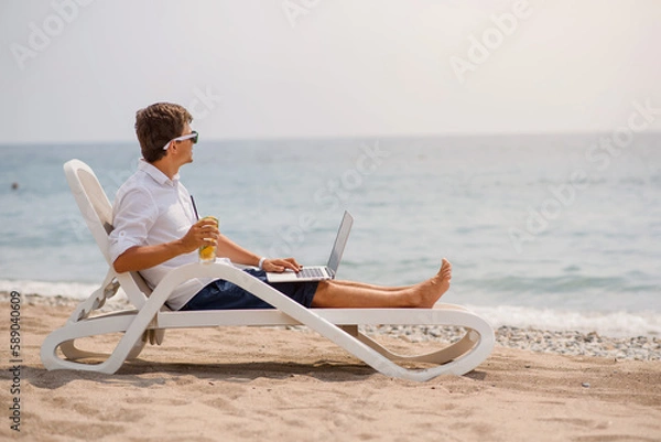 Fototapeta Young man in sunglasses and white shirt is lying on a beach chair on the seaside holding a cocktail and a laptop.