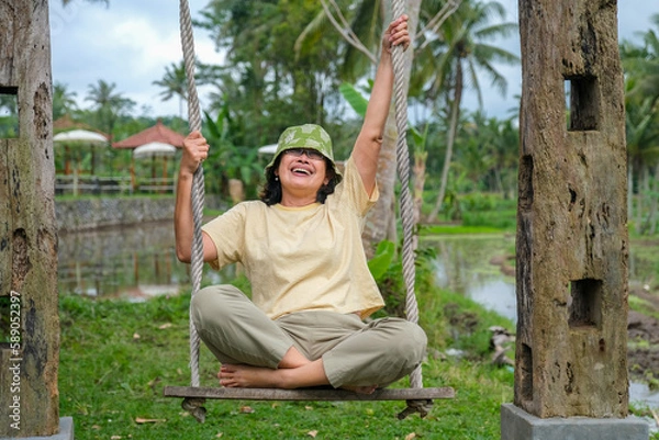 Fototapeta A woman wearing a hat and casual clothes sits on a wooden swing, smiling cheerfully.