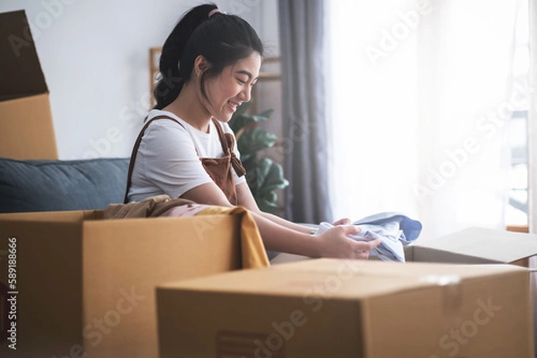Fototapeta New house, asian woman put the clothes in the box while feeling proud and excited about buying a house with a mortgage loan. Young asian woman first time buyers unpacking in dream home or apartment