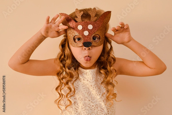 Fototapeta Little cute girl in a carnival masquerade deer mask made of shiny glitter foamiran on a beige background. Happy child in deer costume. The child is preparing for the masquerade.
