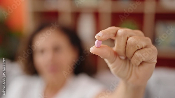 Fototapeta Middle age hispanic woman sitting on sofa holding pill at home