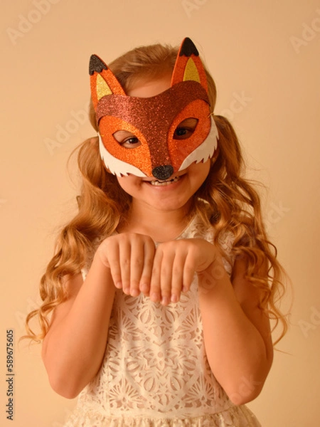 Fototapeta A child in a masquerade carnival mask of a fox, hand-made from glitter foamiran. Little cute cheerful comical girl in the role of a fox. The child is preparing for the carnival, holidays.