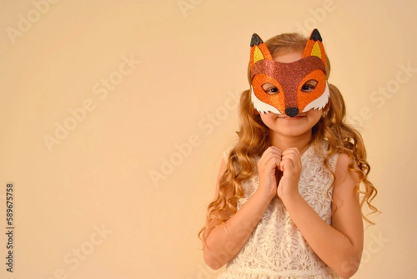 Fototapeta A child in a masquerade carnival mask of a fox, hand-made from glitter foamiran. Little cute cheerful comical girl in the role of a fox. The child is preparing for the carnival, holidays.