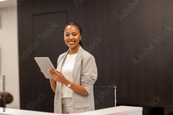 Fototapeta The young African-American female receptionist stands behind the reception desk with a digital tablet in her hands and smiles at the camera.