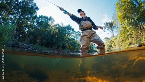 Fototapeta Woman angler on the river. Woman stands in the water in waders and casts the line. Woman fishing on the river