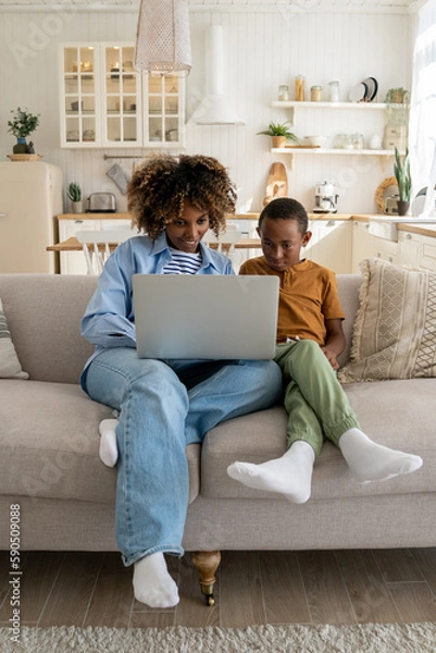 Fototapeta Happy African American family mother and little kid son watching cartoons on laptop together, sitting on couch at home. Sleeping mom and child looking at computer screen having video call