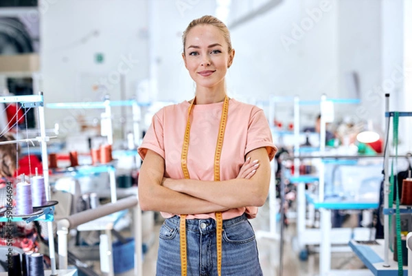 Fototapeta confident woman dressmaker standing in textile factory at workshop, attractive tailor after work stand posing, looking at camera smiling. female use measuring tape for sewing and making garments