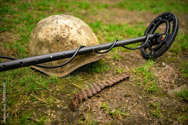 Fototapeta Search for iron finds with a metal detector. Military archaeology.The metal detector lies on an iron helmet in the field.