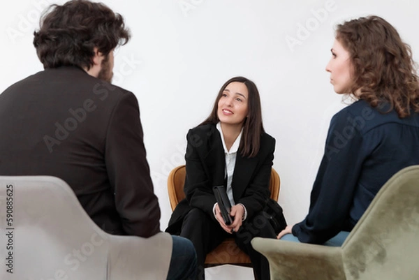 Fototapeta A female psychologist sitting opposite of family couple with a notebook in her hands and attentively listens to her clients at the reception during a psychological counseling