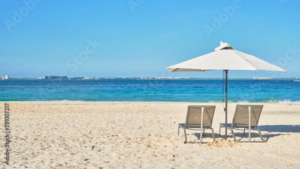 Obraz 2 beach chairs and an a white umbrella on the beaches of cancun on the white sand. in the background a turquoise blue ocean and different hues of blue, small buildings and a sky full of clouds.