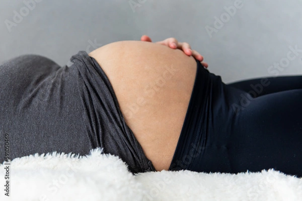 Obraz Pregnant woman lying on her back and resting while putting her hand on her shown belly. Woman relaxing due to pregnancy pain and tiredness. Maternity, pregnancy concept. Side view, Gray background. 