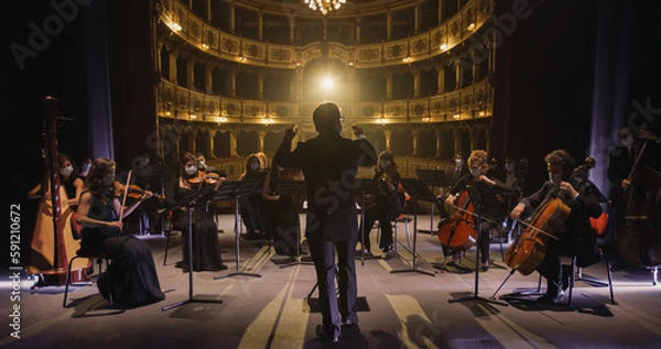 Obraz Back View of Professional Conductor Directing Symphony Orchestra with Performers Wearing Medical Masks, Playing Violins, Cello and Trumpet on Classic Theatre with Curtain Stage During Music Concert