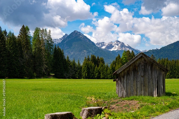 Obraz View of mountain peaks over lush green meadows and forests with shed in foreground on sunny day with blue sky and white clouds