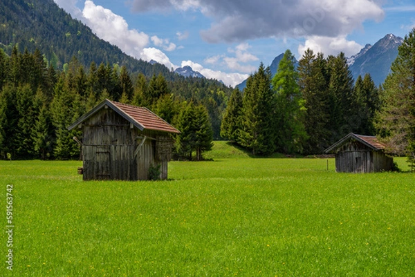 Obraz View to two sheds on green meadow on a sunny day, blue sky with white clouds and mountains in background