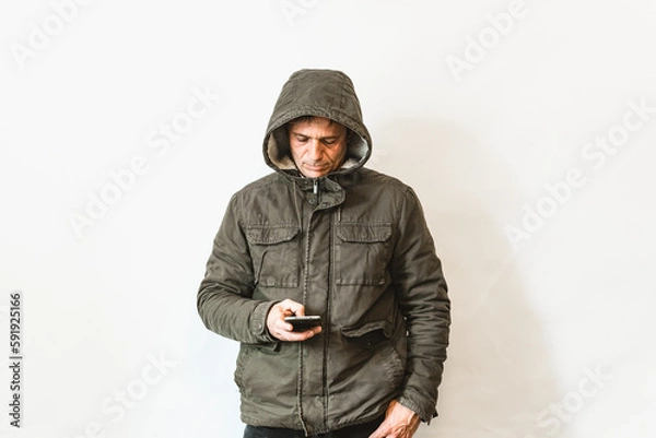 Fototapeta Happy laughing, smiling middle aged senior man wearing green parka standing looking at smartphone isolated over white wall, portrait