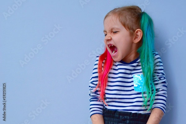 Fototapeta The child is a rebel. A little girl with colored hair and crossed arms on her chest against a gray background stubbornly turns aside, not wanting to listen to anyone. Parenting problems, naughty child