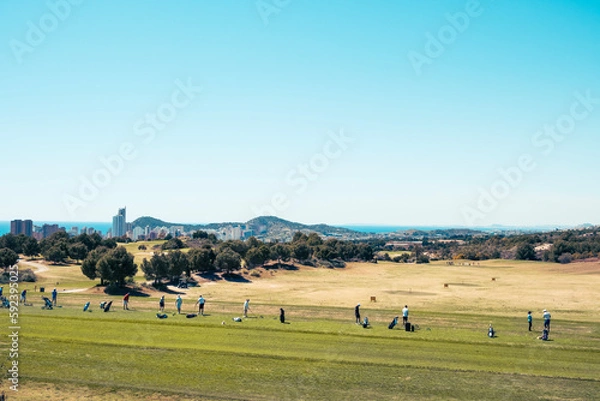Fototapeta Active leisure in Benidorm, Spain. People are playing golf in golf course on sunny day with the skyscrapers of Benidorm and the Mediterranean Sea in the background