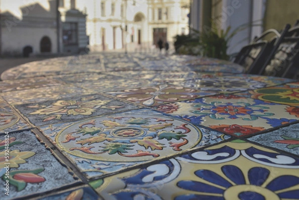 Fototapeta Wet surface of a colorful mosaic table with raindrops in a street cafe against the backdrop of an abstract cityscape.
