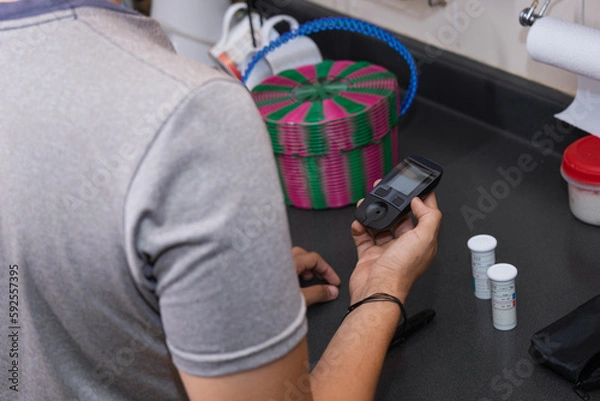Obraz Young Latino man doing blood sugar test in the kitchen. Blood glucose test. Drawing blood for medical test. Using glucose meter in the kitchen.