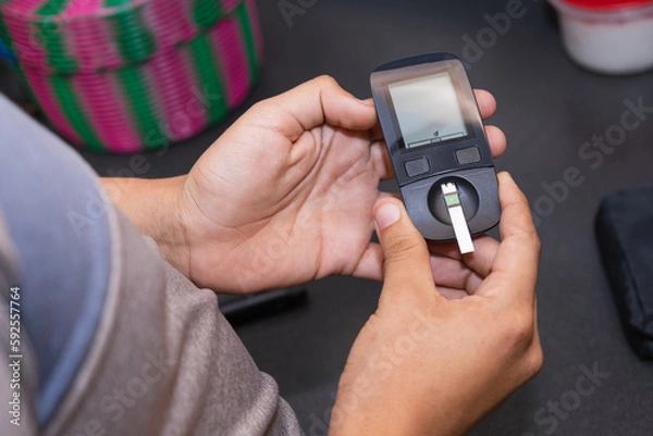 Fototapeta Young Latino man doing blood sugar test in the kitchen. Blood glucose test. Drawing blood for medical test. Using glucose meter in the kitchen.