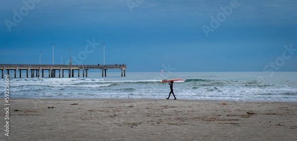 Obraz PORT ARANSAS, TX - 9 FEB 2023: Young surfer carries a surfboard on the beach, near the water of the Gulf of Mexico and a pier, in evening light.