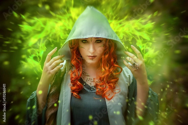 Fototapeta A young woman in a cloak, with a staff and a green dress in the form of a druid. Forest witch with bright red hair. Makeup with black pencil.