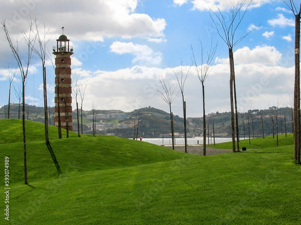 Fototapeta Lisbon, Portugal - Belem Lighthouse, a small red brick and concrete building near the waterfront, surrounded by bare trees under a cloudy blue sky.  Image has copy space.