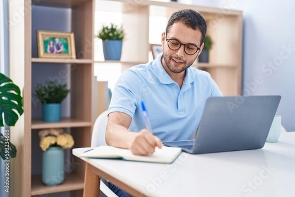 Fototapeta Young man using laptop writing on notebook at home