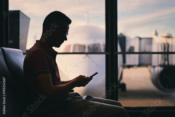 Fototapeta Traveler using phone while waiting for flight. Silhouette of man waiting by window of airport terminal behind which is airplane..