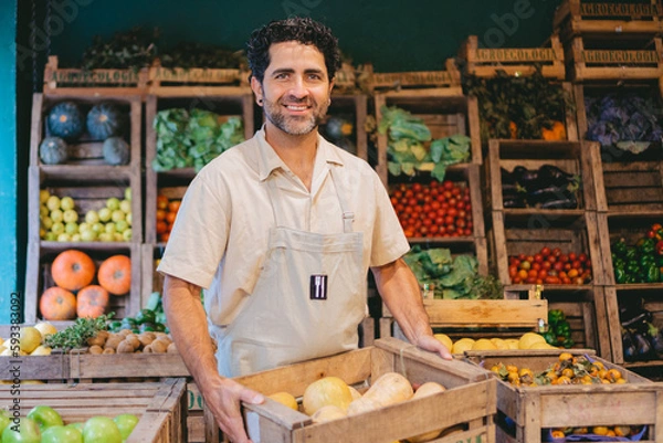 Obraz Middle-aged Latin man with a smile holding a crate of squash in a greengrocer's shop while looking at the camera.