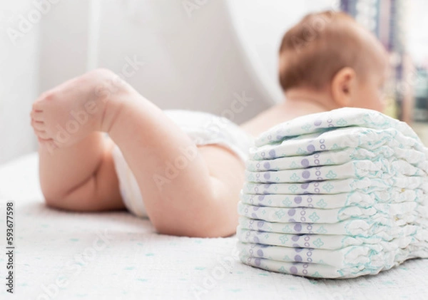 Fototapeta The legs of a baby in a diaper against the background of a stack of children's comfortable diapers, close-up