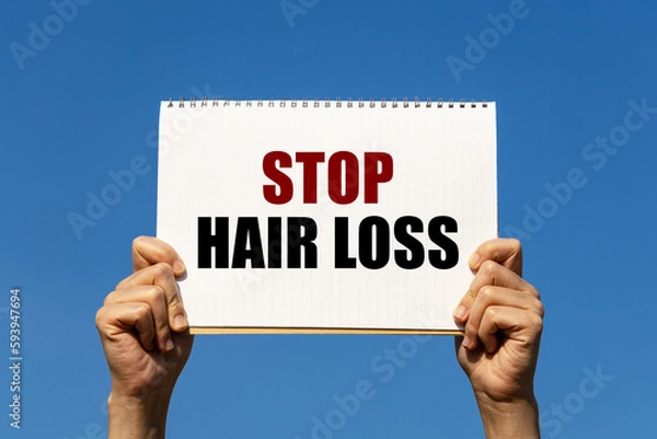 Fototapeta Stop hair loss text on notebook paper held by 2 hands with isolated blue sky background. This message can be used as business concept about stop hair loss.