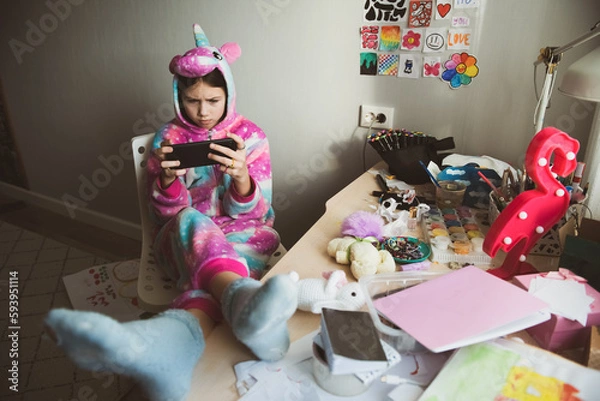 Fototapeta Serious teen girl using phone, playing phone games in messy childrens room, kid playing among the many toys at home, a lot of toys and things scattered on table, dirty home