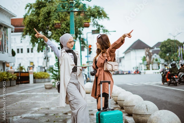Fototapeta two female travellers pointing to the opposite way while standing together at the sidewalk bring their travel equipment